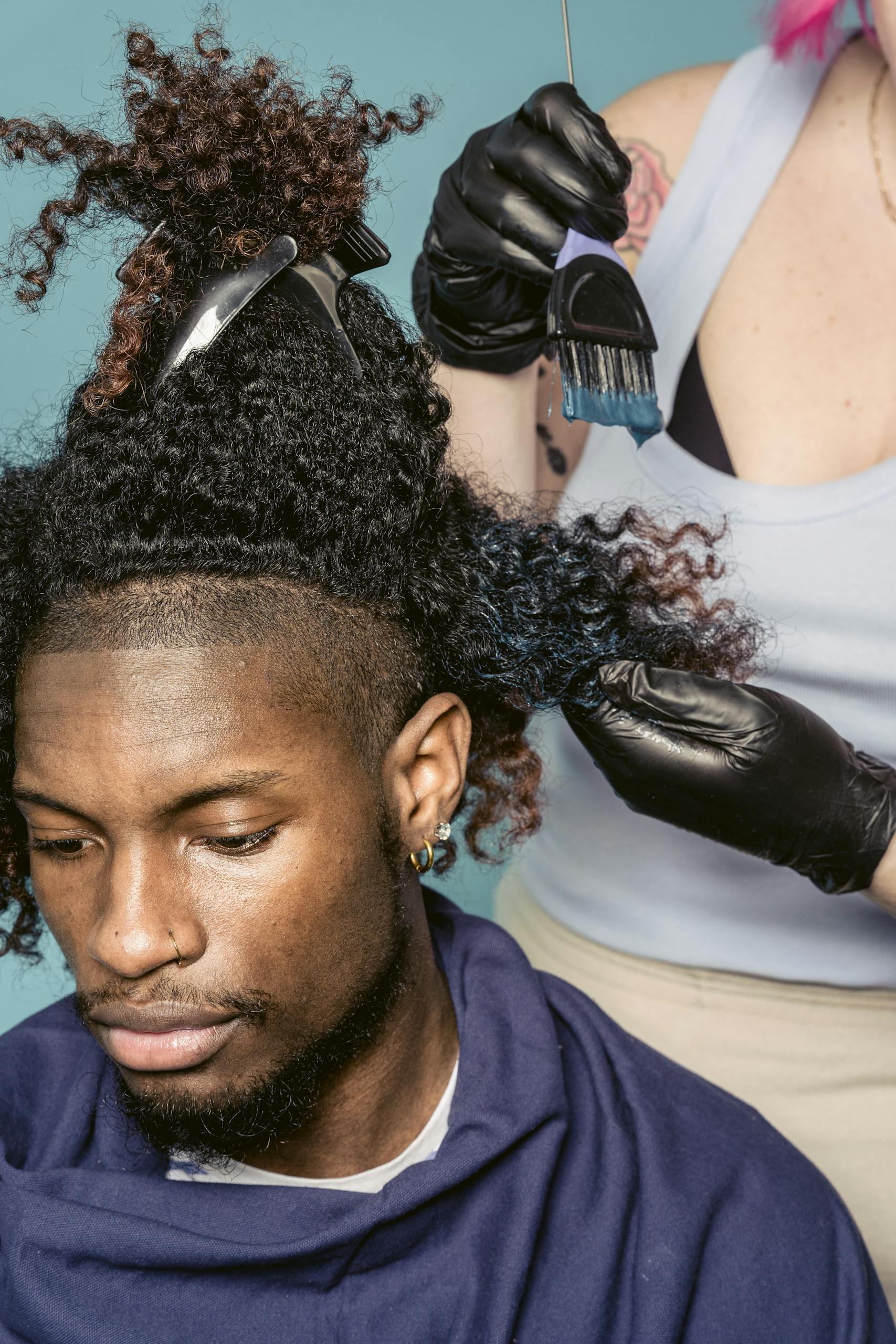 A hairstylist wearing gloves applies blue dye to a man's curly hair in a salon.